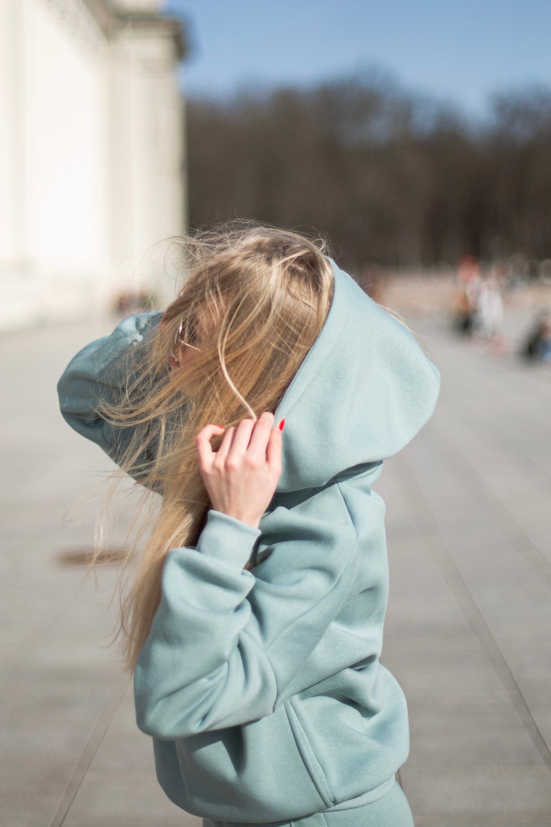 K&ouml;nnte beinhalten: Ein hellblauer Hoodie mit passendem Trainingsanzug. Die Person tr&auml;gt die Kapuze &uuml;ber dem Kopf. Die Person hat lange blonde Haare und roten Nagellack. Der Hintergrund ist eine verschwommene Szene im Freien.