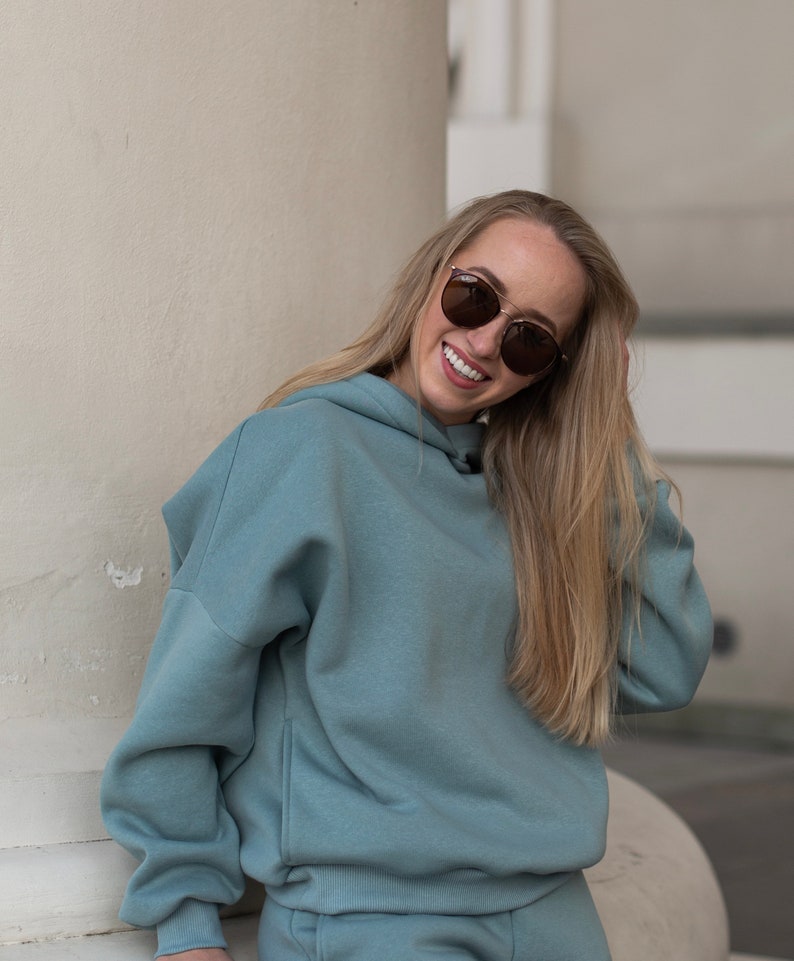 K&ouml;nnte beinhalten: Hellblauer Hoodie und passendes Jogginghosen-Set. Der Hoodie hat eine Kapuze mit Kordelzug und eine K&auml;ngurutasche. Die Person tr&auml;gt eine Sonnenbrille mit goldenem Rahmen. Das Outfit ist aus weichem, bequemem Material.