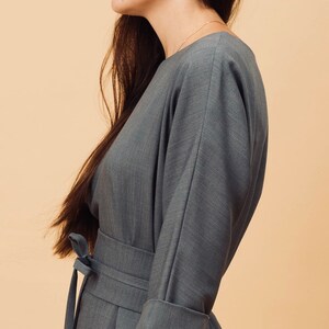 May include: A grey dress with a wrap-around waist and three-quarter sleeves. The dress has a tie belt and cuffed sleeves. The model is wearing a gold necklace and small earrings. The background is a solid beige colour.