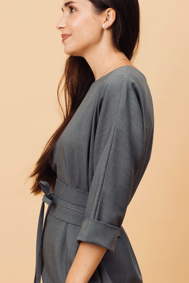 May include: A grey dress with a wrap-around waist and three-quarter sleeves. The dress has a tie belt and cuffed sleeves. The model is wearing a gold necklace and small earrings. The background is a solid beige colour.