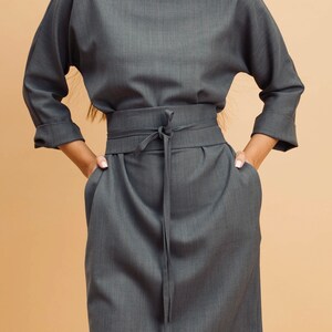 May include: A grey dress with a round neckline, three-quarter sleeves, and a tie-waist belt. The dress has a knee-length skirt and pockets. The fabric has a subtle texture. The model is wearing a gold necklace.