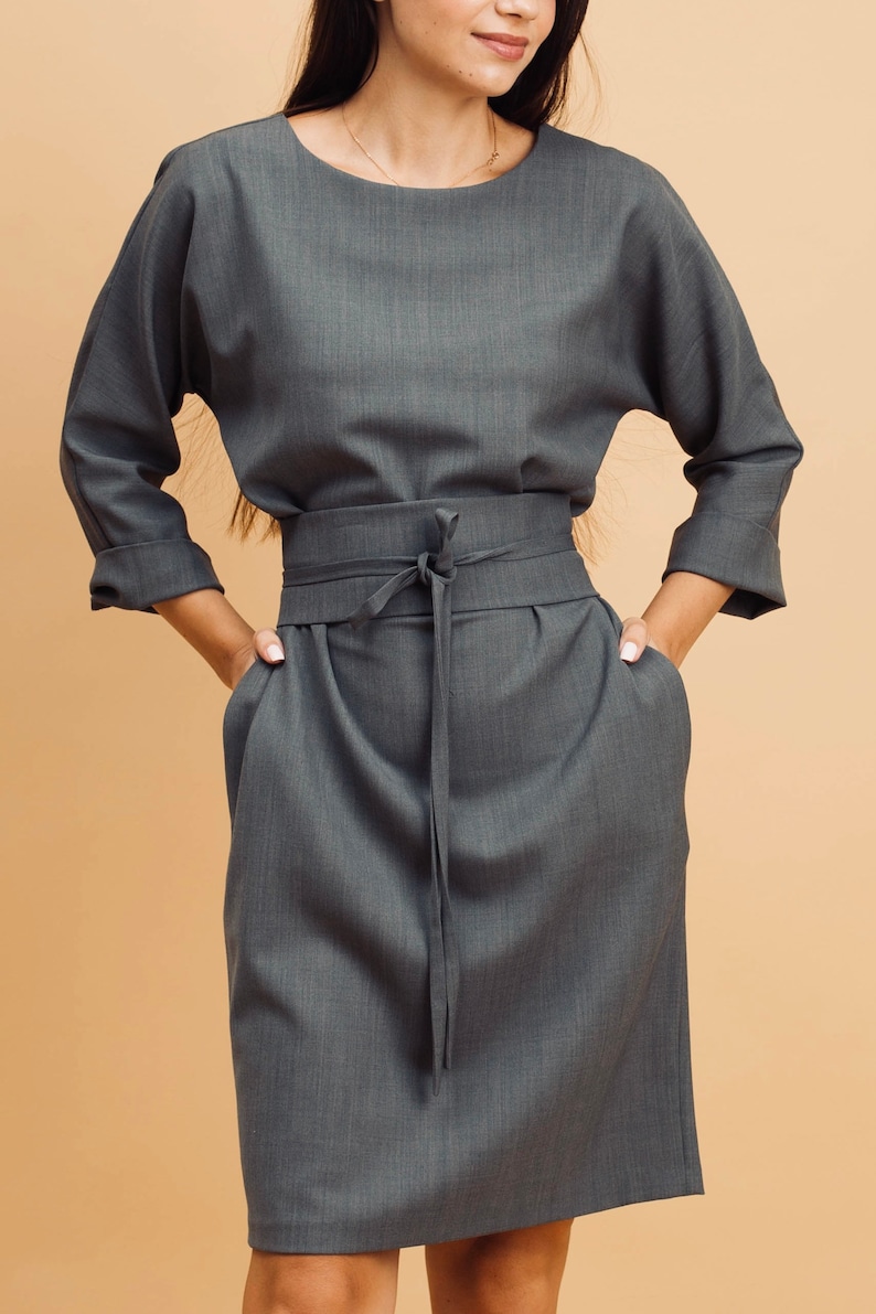 May include: A grey dress with a round neckline, three-quarter sleeves, and a tie-waist belt. The dress has a knee-length skirt and pockets. The fabric has a subtle texture. The model is wearing a gold necklace.
