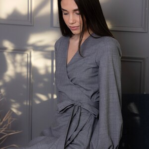 May include: A gray wrap dress with long sleeves and a tie at the waist. The dress has a V-neckline and a tailored fit. The sleeves are long and have a slight puff at the shoulder. The dress is made of a soft, woven fabric.