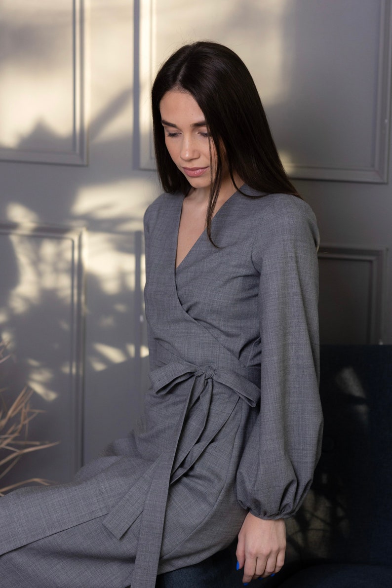 May include: A gray wrap dress with long sleeves and a tie at the waist. The dress has a V-neckline and a tailored fit. The sleeves are long and have a slight puff at the shoulder. The dress is made of a soft, woven fabric.