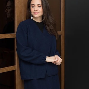 Op de afbeelding: Een marineblauw tweedelig outfit, bestaande uit een jas met lange mouwen en een bijpassende rok. De jas heeft een V-hals en de rok is knielang. De outfit is gemaakt van een zachte, getextureerde stof. De vrouw draagt een zwarte top onder de jas.