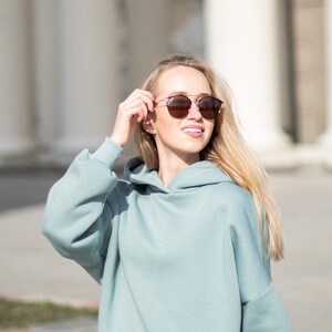 K&ouml;nnte beinhalten: Eine Frau tr&auml;gt einen hellblauen Kapuzenpullover und eine passende Jogginghose sowie eine runde Sonnenbrille. Die Sonnenbrille hat einen Doppelsteg und braune Gl&auml;ser. Die Frau l&auml;chelt und h&auml;lt die Sonnenbrille. Im Hintergrund wei&szlig;e S&auml;ulen.