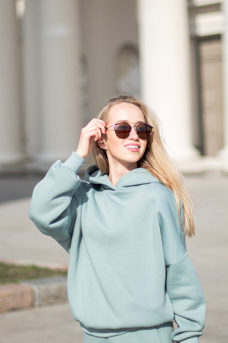 K&ouml;nnte beinhalten: Eine Frau tr&auml;gt einen hellblauen Kapuzenpullover und eine passende Jogginghose sowie eine runde Sonnenbrille. Die Sonnenbrille hat einen Doppelsteg und braune Gl&auml;ser. Die Frau l&auml;chelt und h&auml;lt die Sonnenbrille. Im Hintergrund wei&szlig;e S&auml;ulen.