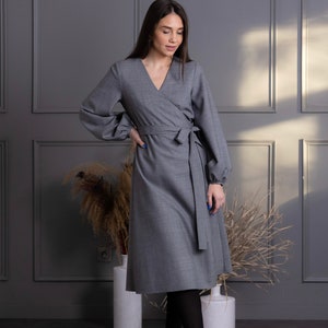 May include: A gray wrap dress with long sleeves and a V-neck. The dress features a tie at the waist and is paired with black tights and heels. The dress is knee-length and made of a solid gray fabric.