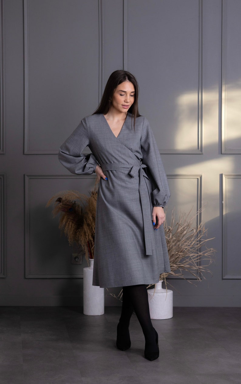 May include: A gray wrap dress with long sleeves and a V-neck. The dress features a tie at the waist and is paired with black tights and heels. The dress is knee-length and made of a solid gray fabric.