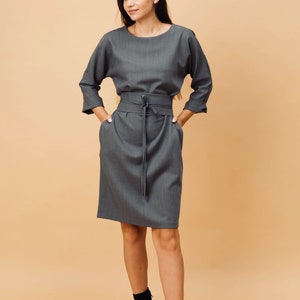 May include: A grey dress with a matching belt tied at the waist. The dress features three-quarter sleeves and pockets. The model is wearing black ankle boots. The background is a solid beige colour.