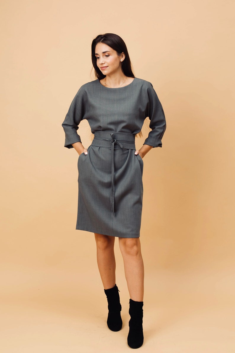 May include: A grey dress with a matching belt tied at the waist. The dress features three-quarter sleeves and pockets. The model is wearing black ankle boots. The background is a solid beige colour.