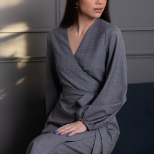 May include: A gray wrap dress with long sleeves and a V-neck. The dress features a tie at the waist and is made of a textured fabric. The model is wearing gold earrings and has blue nail polish. The dress is suitable for formal or business wear.