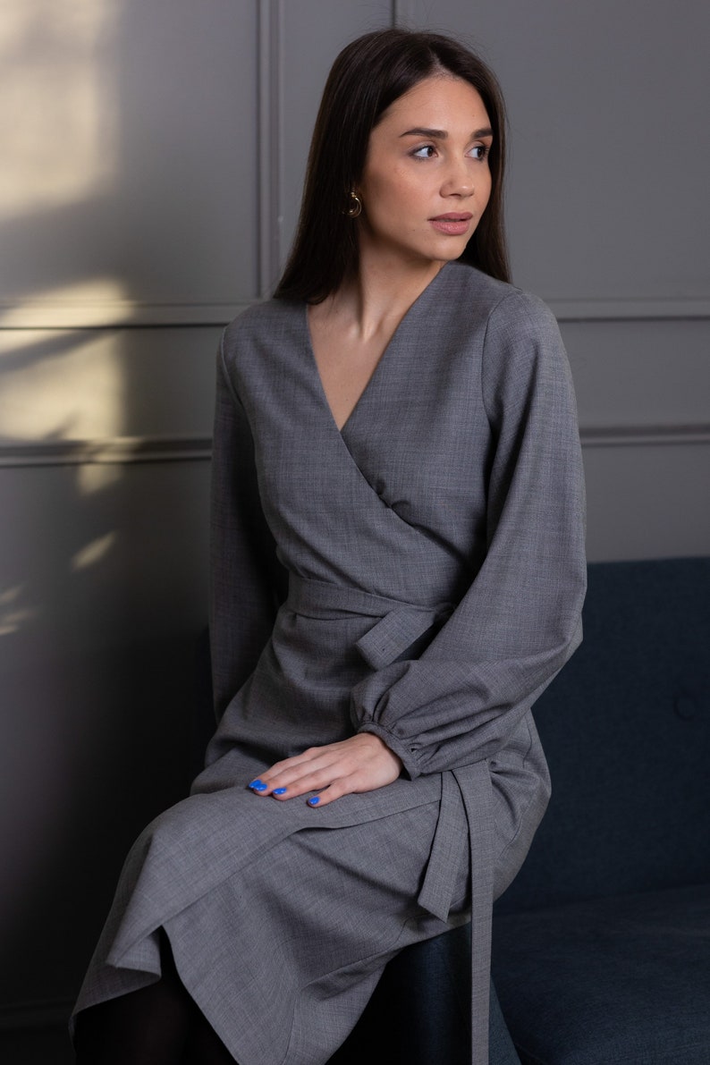 May include: A gray wrap dress with long sleeves and a V-neck. The dress features a tie at the waist and is made of a textured fabric. The model is wearing gold earrings and has blue nail polish. The dress is suitable for formal or business wear.