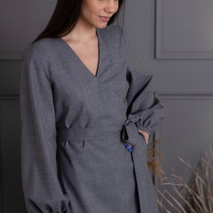 May include: A gray wrap dress with long sleeves and a V-neck. The dress features a tie at the waist and a blue button detail. The sleeves have a slight puff at the shoulders. The dress is made of a textured fabric.