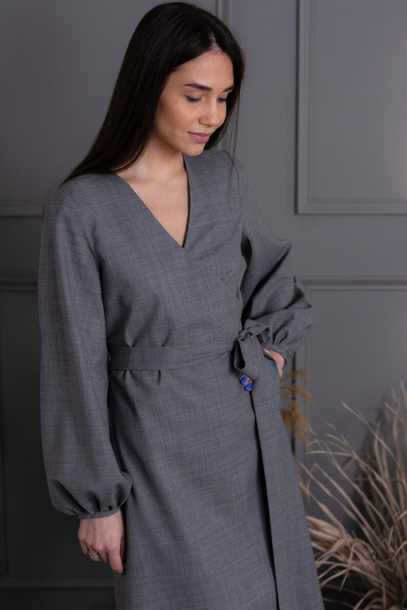May include: A gray wrap dress with long sleeves and a V-neck. The dress features a tie at the waist and a blue button detail. The sleeves have a slight puff at the shoulders. The dress is made of a textured fabric.