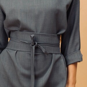 May include: A grey two-piece outfit. The top has three-quarter sleeves, and the trousers have a matching fabric belt tied at the waist. The fabric has a subtle texture. The outfit is a neutral colour.