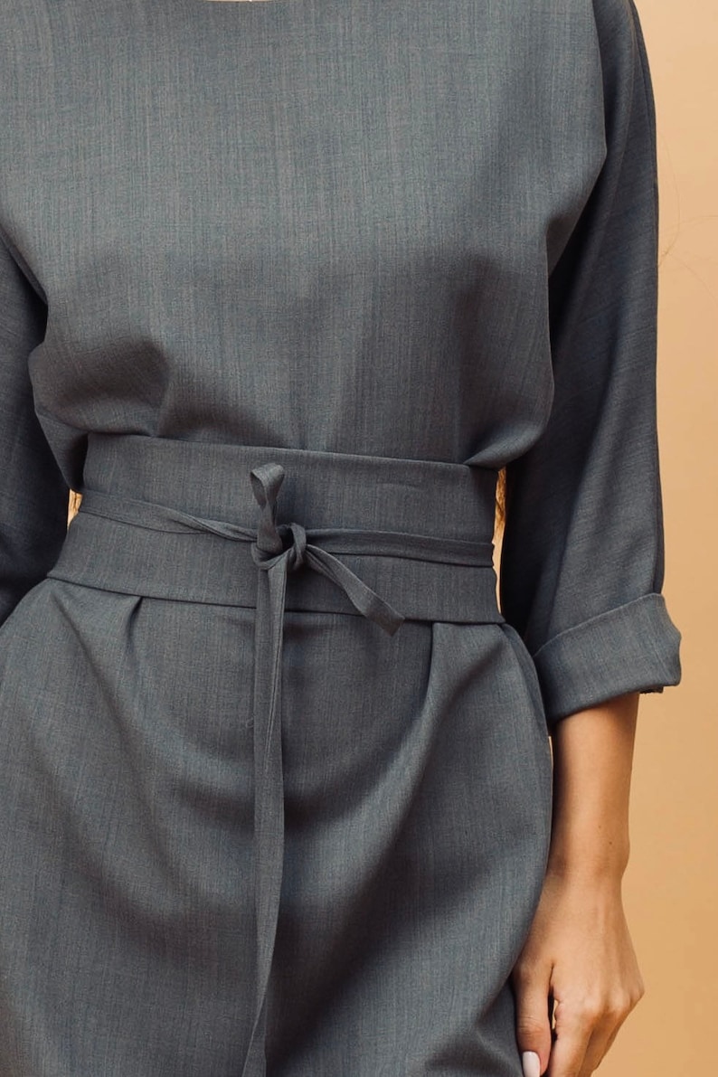 May include: A grey two-piece outfit. The top has three-quarter sleeves, and the trousers have a matching fabric belt tied at the waist. The fabric has a subtle texture. The outfit is a neutral colour.