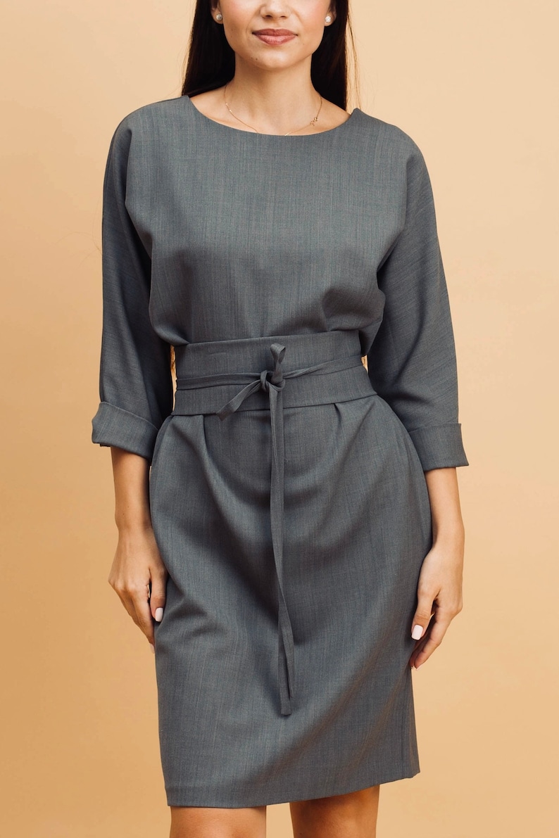 May include: A grey dress with a boat neck and three-quarter sleeves. The dress has a tie-waist detail and a knee-length skirt. The fabric has a subtle texture, and the overall style is simple and elegant.