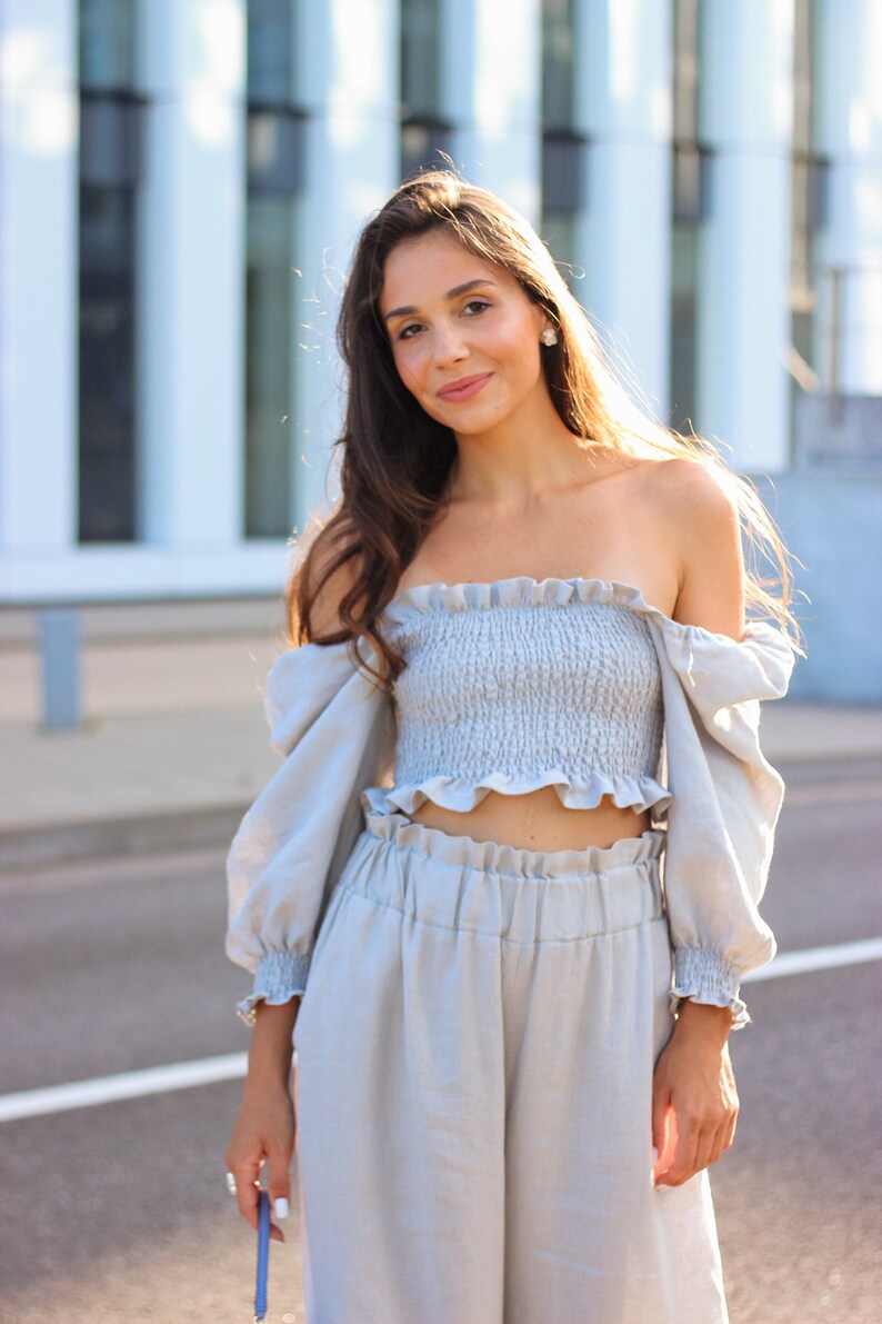 K&ouml;nnte beinhalten: Hellblaues zweiteiliges Outfit, bestehend aus einem schulterfreien Crop-Top mit R&uuml;schen und einer Hose mit weitem Bein. Das Oberteil hat lange, gepuffte &Auml;rmel. Das Outfit ist aus leichtem Stoff gefertigt, ideal f&uuml;r warmes Wetter.