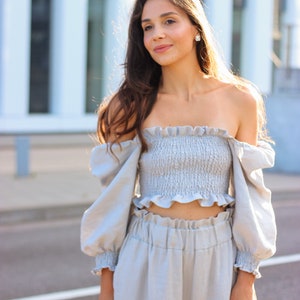 K&ouml;nnte beinhalten: Hellblaues zweiteiliges Outfit mit einem ger&uuml;schten schulterfreien Crop-Top und einer passenden hoch taillierten Hose. Das Outfit hat lange, puff&auml;rmelige &Auml;rmel und ist aus einem weichen, flie&szlig;enden Stoff gefertigt. Das Model tr&auml;gt das Outfit im Freien.
