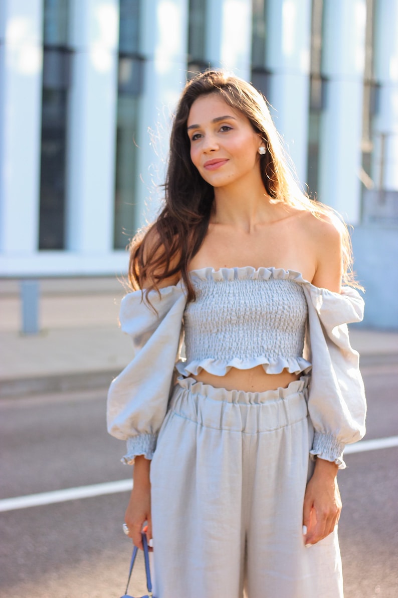K&ouml;nnte beinhalten: Hellblaues zweiteiliges Outfit mit einem ger&uuml;schten schulterfreien Crop-Top und einer passenden hoch taillierten Hose. Das Outfit hat lange, puff&auml;rmelige &Auml;rmel und ist aus einem weichen, flie&szlig;enden Stoff gefertigt. Das Model tr&auml;gt das Outfit im Freien.