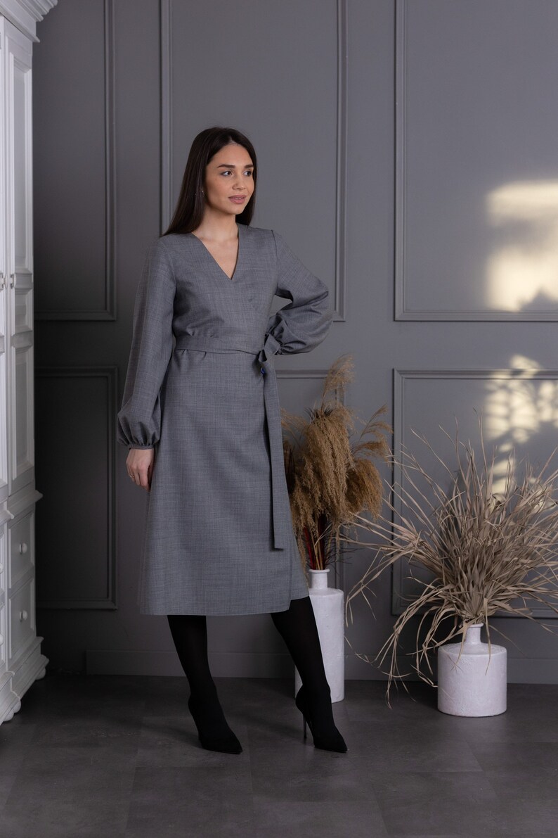 May include: A gray wrap dress with long sleeves and a V-neck. The dress features a tie belt at the waist and is paired with black tights and high-heeled boots. The dress is made of a textured fabric.