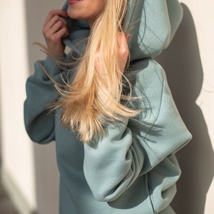 K&ouml;nnte beinhalten: Hellblauer Kapuzenpullover mit passender Jogginghose. Der Kapuzenpullover hat eine gro&szlig;e Kapuze und lange &Auml;rmel. Das Model tr&auml;gt eine Sonnenbrille und hat blondes Haar. Das Outfit ist aus einem weichen, bequemen Material.
