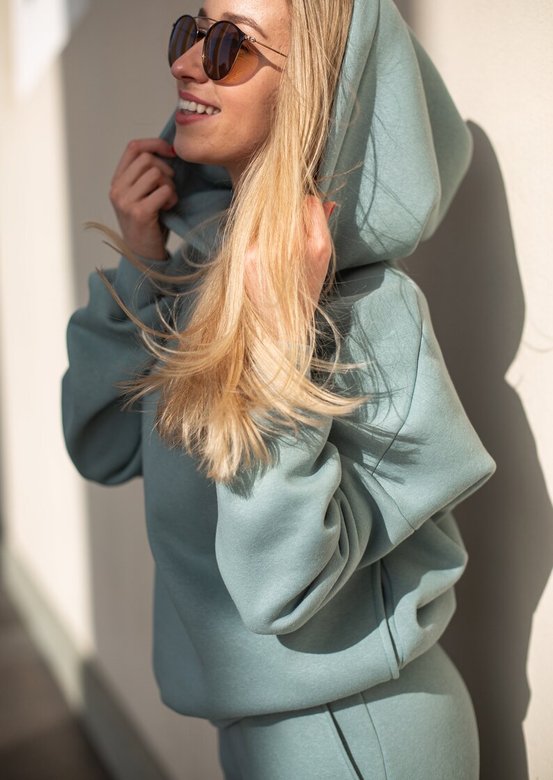 K&ouml;nnte beinhalten: Hellblauer Kapuzenpullover mit passender Jogginghose. Der Kapuzenpullover hat eine gro&szlig;e Kapuze und lange &Auml;rmel. Das Model tr&auml;gt eine Sonnenbrille und hat blondes Haar. Das Outfit ist aus einem weichen, bequemen Material.