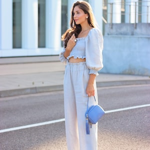 K&ouml;nnte beinhalten: Hellblaues Crop-Top mit Puff&auml;rmeln und passender hochtaillierter Hose. Eine kleine, quadratische, hellblaue Handtasche mit Quaste wird gehalten. Das Outfit wird durch hellblaue Sandalen erg&auml;nzt. Die Kleidung ist aus leinenartigem Material.