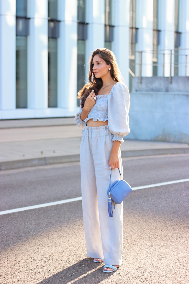 K&ouml;nnte beinhalten: Hellblaues Crop-Top mit Puff&auml;rmeln und passender hochtaillierter Hose. Eine kleine, quadratische, hellblaue Handtasche mit Quaste wird gehalten. Das Outfit wird durch hellblaue Sandalen erg&auml;nzt. Die Kleidung ist aus leinenartigem Material.