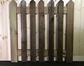May include: A wooden picket fence gate with pointed tops. The gate is made of brown stained wood and is approximately 90 cm tall.
