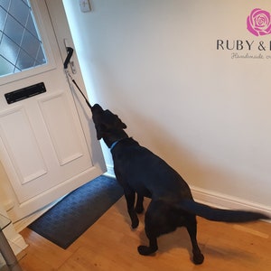 May include: A black dog is pulling on a black lead attached to a door handle. The dog is looking at the door. The door is white and has a glass panel with a diamond pattern. The dog is wearing a blue collar. The text "RUBY & ROSE Handmade creations" is visible on the wall.