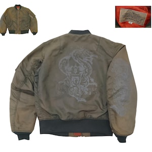 Puede incluir: Chaqueta bomber verde oliva con un dise&ntilde;o de calavera y serpiente en la espalda. La chaqueta tiene cierre de cremallera, pu&ntilde;os de canal&eacute; y un bolsillo en la manga. El forro interior es naranja. La etiqueta dice AVIREX LTD.