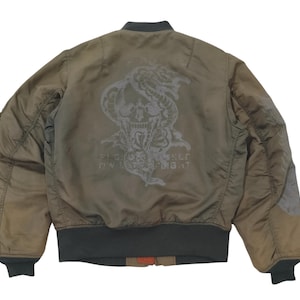 Puede incluir: Chaqueta bomber verde oliva con un dise&ntilde;o de calavera y serpiente en la espalda. La chaqueta tiene cuello, pu&ntilde;os y dobladillo de canal&eacute;. El gr&aacute;fico incluye el texto "Fight or Flight".