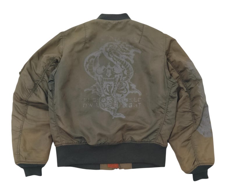Puede incluir: Chaqueta bomber verde oliva con un dise&ntilde;o de calavera y serpiente en la espalda. La chaqueta tiene cuello, pu&ntilde;os y dobladillo de canal&eacute;. El gr&aacute;fico incluye el texto "Fight or Flight".