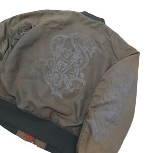 Puede incluir: Chaqueta bomber verde oliva con un dise&ntilde;o de calavera y serpiente gris oscuro en la espalda y un drag&oacute;n en la manga. La chaqueta tiene cuello, pu&ntilde;os y dobladillo de canal&eacute; verde oscuro. El texto "BIG WILD FLIGHT" es visible.