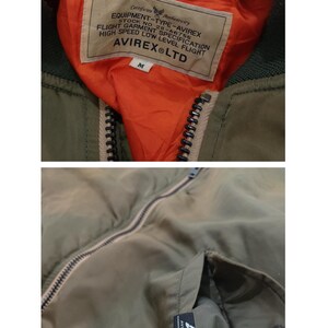 Puede incluir: Chaqueta bomber verde oliva con forro naranja. La chaqueta tiene cierre de cremallera y cuello verde oscuro. La etiqueta interior dice "EQUIPMENT-TYPE-AVIREX" y "AVIREX LTD". Una etiqueta con texto es visible dentro del bolsillo.