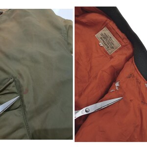 Puede incluir: Chaqueta bomber verde oliva con cuello negro y forro naranja. La chaqueta tiene cierre de cremallera y un bolsillo en la parte delantera. El interior presenta una etiqueta con texto. La chaqueta se muestra con unas tijeras.