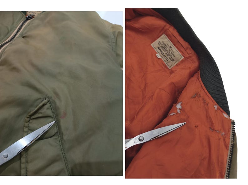 Puede incluir: Chaqueta bomber verde oliva con cuello negro y forro naranja. La chaqueta tiene cierre de cremallera y un bolsillo en la parte delantera. El interior presenta una etiqueta con texto. La chaqueta se muestra con unas tijeras.
