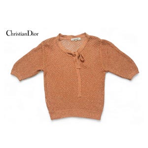 Puede incluir: Un suéter de punto marrón de tejido abierto con un lazo en el cuello. El suéter tiene mangas cortas y cuello en V. La etiqueta dice "Christian Dior".