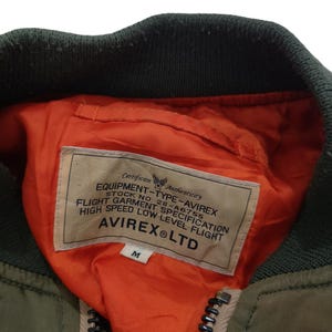 Puede incluir: Primer plano de una chaqueta bomber verde oliva con cuello verde oscuro y forro naranja. La chaqueta tiene una cremallera y una etiqueta con el texto "AVIREX&reg; LTD". La etiqueta tambi&eacute;n incluye el texto "EQUIPMENT-TYPE-AVIREX" y "FLIGHT GARMENT SPECIFICATION".