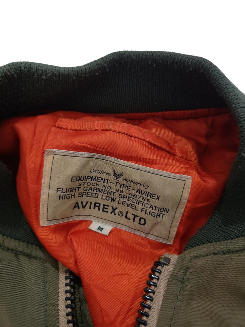 Puede incluir: Primer plano de una chaqueta bomber verde oliva con cuello verde oscuro y forro naranja. La chaqueta tiene una cremallera y una etiqueta con el texto "AVIREX&reg; LTD". La etiqueta tambi&eacute;n incluye el texto "EQUIPMENT-TYPE-AVIREX" y "FLIGHT GARMENT SPECIFICATION".