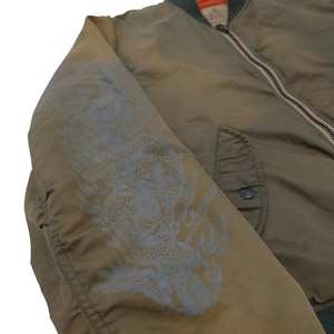 Puede incluir: Chaqueta bomber verde oliva con un dise&ntilde;o de drag&oacute;n en la manga izquierda. La chaqueta tiene cierre de cremallera, cuello verde oscuro y un bolsillo en la parte delantera. El dise&ntilde;o del drag&oacute;n es gris claro.