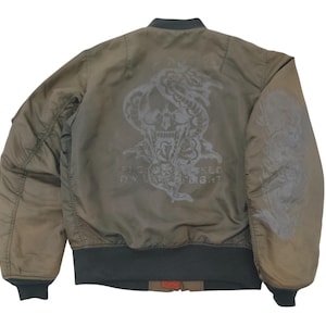 Puede incluir: Chaqueta bomber verde oliva con un gr&aacute;fico de calavera y serpiente en la espalda. La chaqueta tiene cuello, pu&ntilde;os y dobladillo de canal&eacute;. El gr&aacute;fico incluye el texto "FLY LIKE A FIGHT".