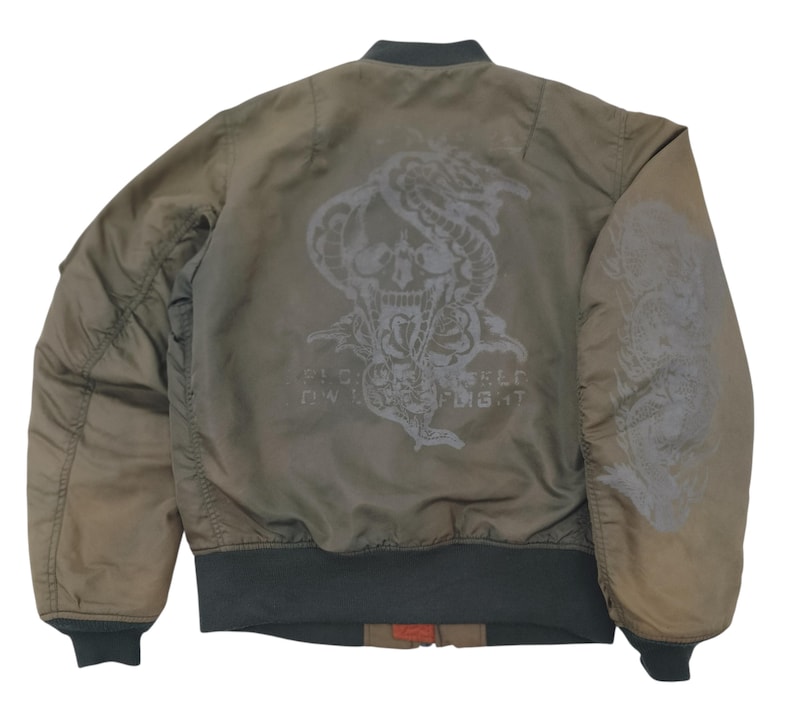 Puede incluir: Chaqueta bomber verde oliva con un gr&aacute;fico de calavera y serpiente en la espalda. La chaqueta tiene cuello, pu&ntilde;os y dobladillo de canal&eacute;. El gr&aacute;fico incluye el texto "FLY LIKE A FIGHT".