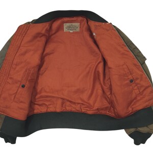 Puede incluir: Chaqueta bomber verde oliva con cuello y pu&ntilde;os negros. El interior es de color naranja vibrante con una textura acolchada. Una peque&ntilde;a etiqueta es visible cerca del cuello. La chaqueta tiene dos bolsillos y cierre de cremallera.