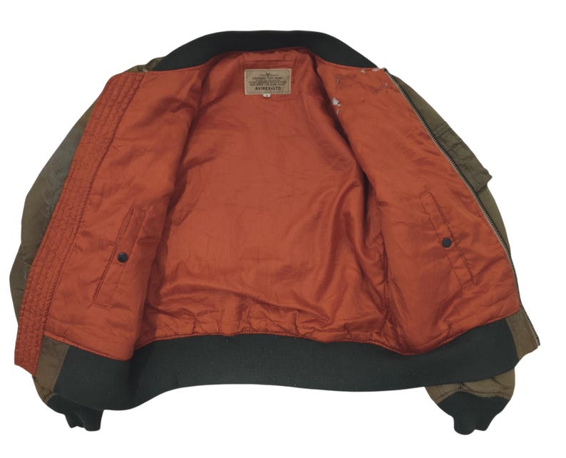Puede incluir: Chaqueta bomber verde oliva con cuello y pu&ntilde;os negros. El interior es de color naranja vibrante con una textura acolchada. Una peque&ntilde;a etiqueta es visible cerca del cuello. La chaqueta tiene dos bolsillos y cierre de cremallera.