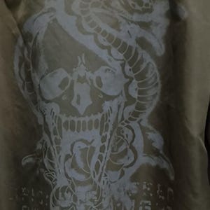 Puede incluir: Chaqueta bomber verde oliva con un gran gr&aacute;fico blanco desgastado en la espalda. El gr&aacute;fico representa una calavera entrelazada con una serpiente y rosas, con texto debajo. La chaqueta tiene un cuello verde.