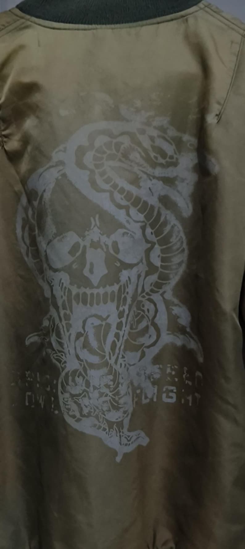 Puede incluir: Chaqueta bomber verde oliva con un gran gr&aacute;fico blanco desgastado en la espalda. El gr&aacute;fico representa una calavera entrelazada con una serpiente y rosas, con texto debajo. La chaqueta tiene un cuello verde.