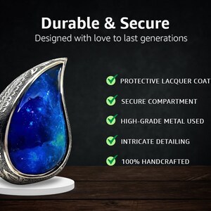 Op de afbeelding: Een zilveren urn in de vorm van een traan met een blauw en wit galaxieontwerp. De urn is handgemaakt en heeft ingewikkelde details. Het is ontworpen om duurzaam en veilig te zijn, met een beschermende lakcoating en een veilig compartiment.